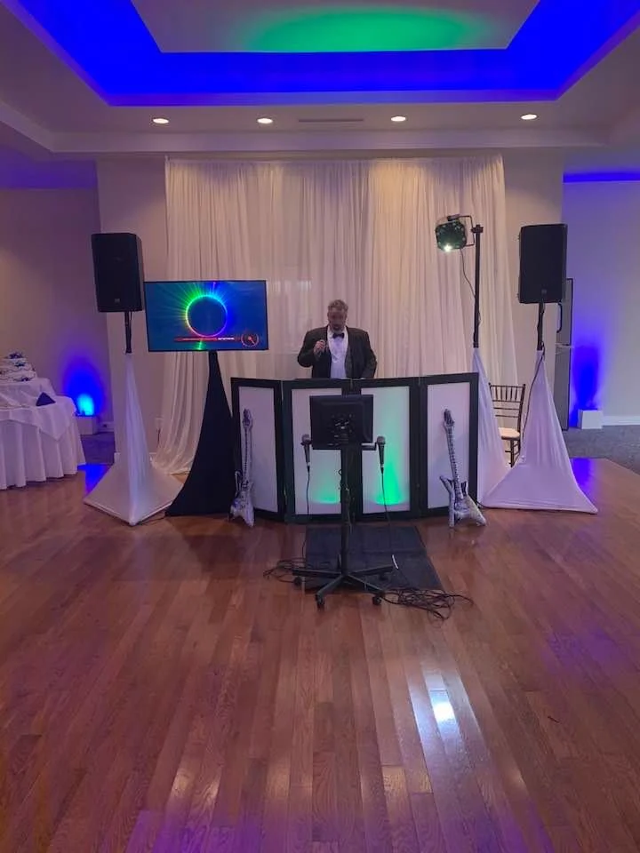 DJ Dano at a white DJ booth in a ballroom with blue and green uplighting