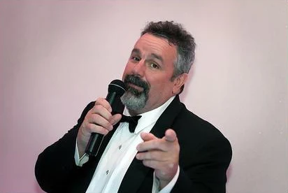 DJ Dano in a tuxedo hosting an event with a microphone